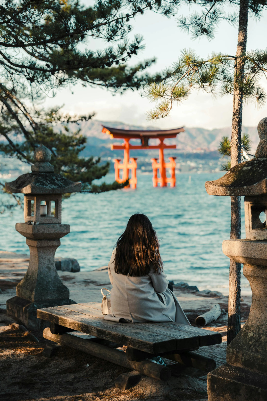 Woman sitting by the sea facing the floating torii gate of Itsukushima Shrine in Miyajima, Japan.